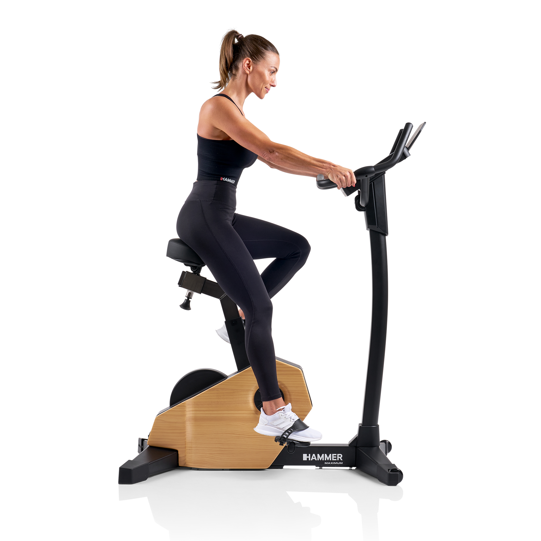 Frau auf dem Heimtrainer. Sie trägt schwarze Sportkleidung. Das Fahrrad ist schwarz und holzfarben.