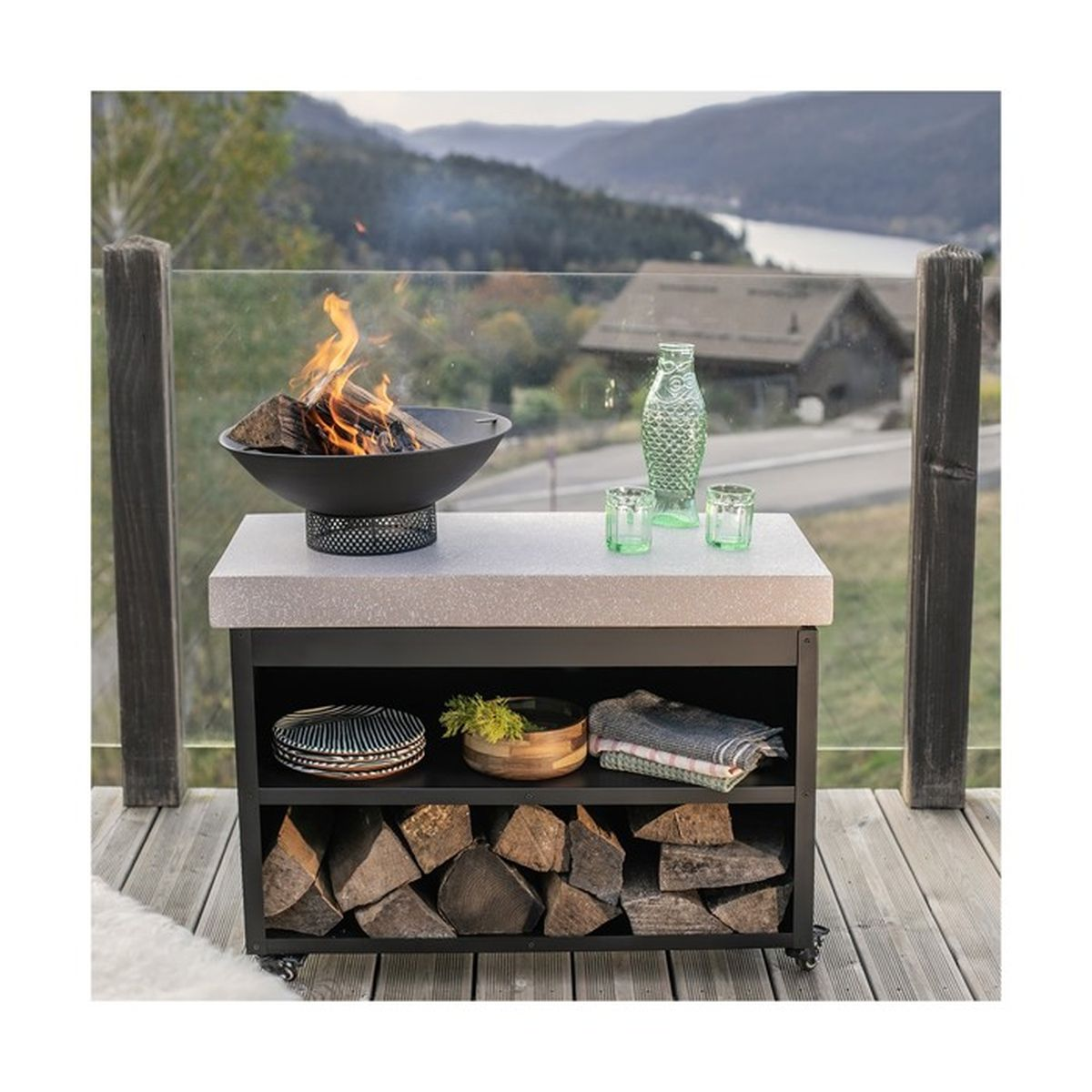 Ein Feuer mit brennendem Holz steht auf einem Tisch mit Regalen für Holz, Geschirr und Handtücher. Im Hintergrund sind ein See und Berge zu sehen.