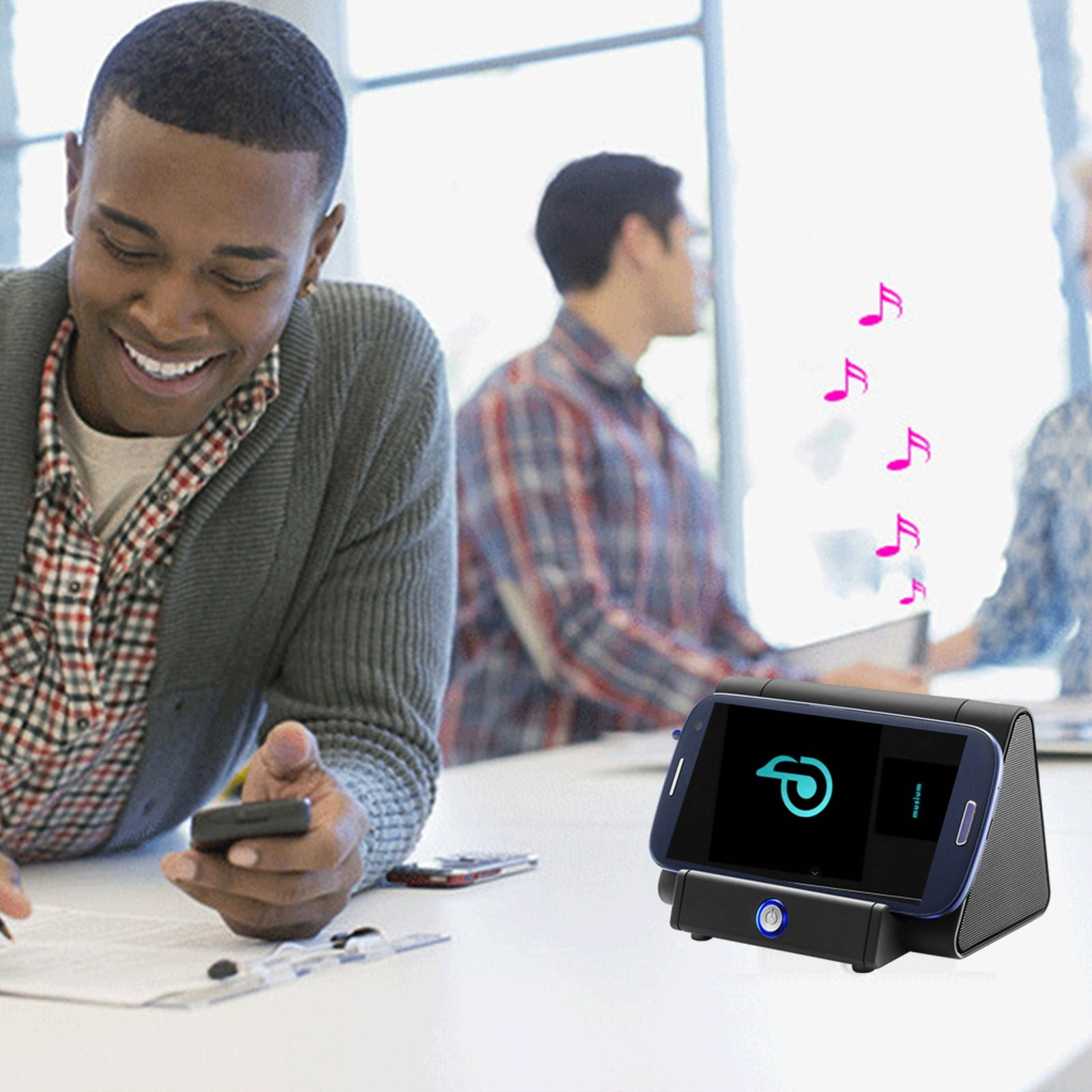 Ein Mann lächelt, während er ein Telefon benutzt. Ein Telefon ist an einem Lautsprecher angedockt, Noten schweben auf. Zwei andere Personen sind im Hintergrund.