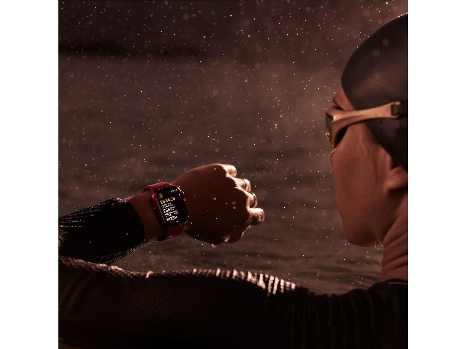 A swimmer looks at their watch displaying workout data. The watch is red, the swimmer is wearing goggles. Water droplets are visible.