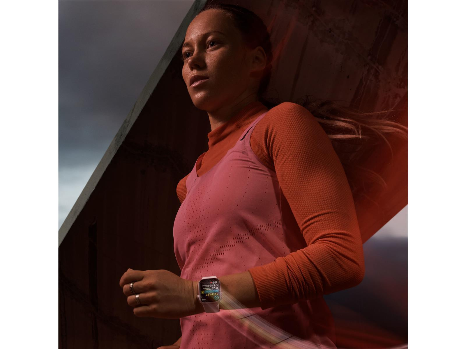 Woman running, wearing pink and orange athletic wear, with a smartwatch. Concrete structure in background.