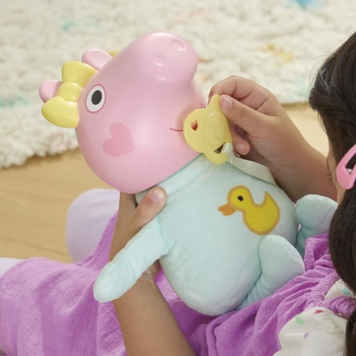Ein Kind hält ein rosa Spielzeugschwein mit einem gelben Schnuller. Das Schwein hat ein Herz und eine Ente auf dem Körper. Ein Mädchen mit rosa Haarspange.