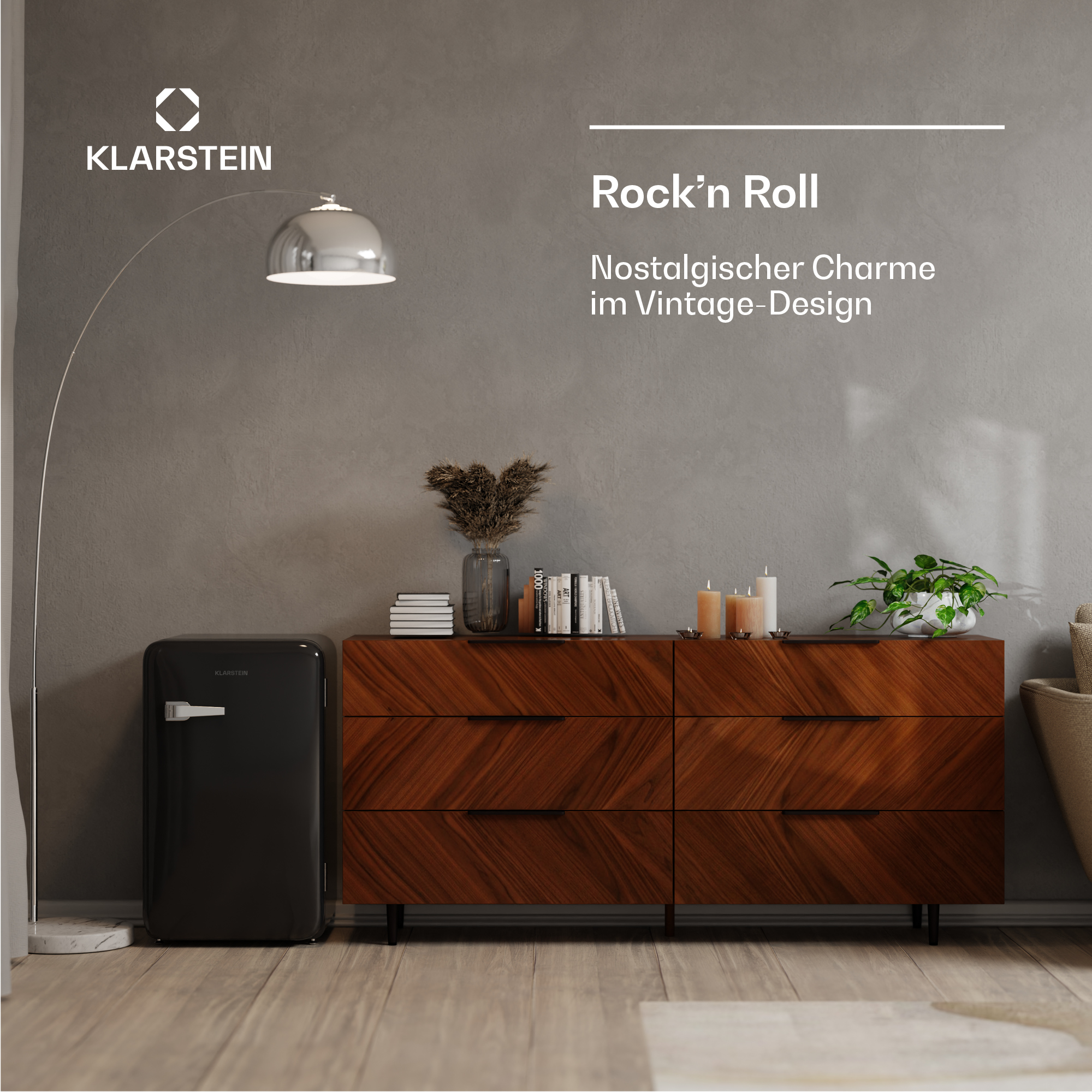 Scena di soggiorno con frigorifero nero, cassettiera in legno, lampada da terra ed elementi decorativi. Sfondo muro grigio.