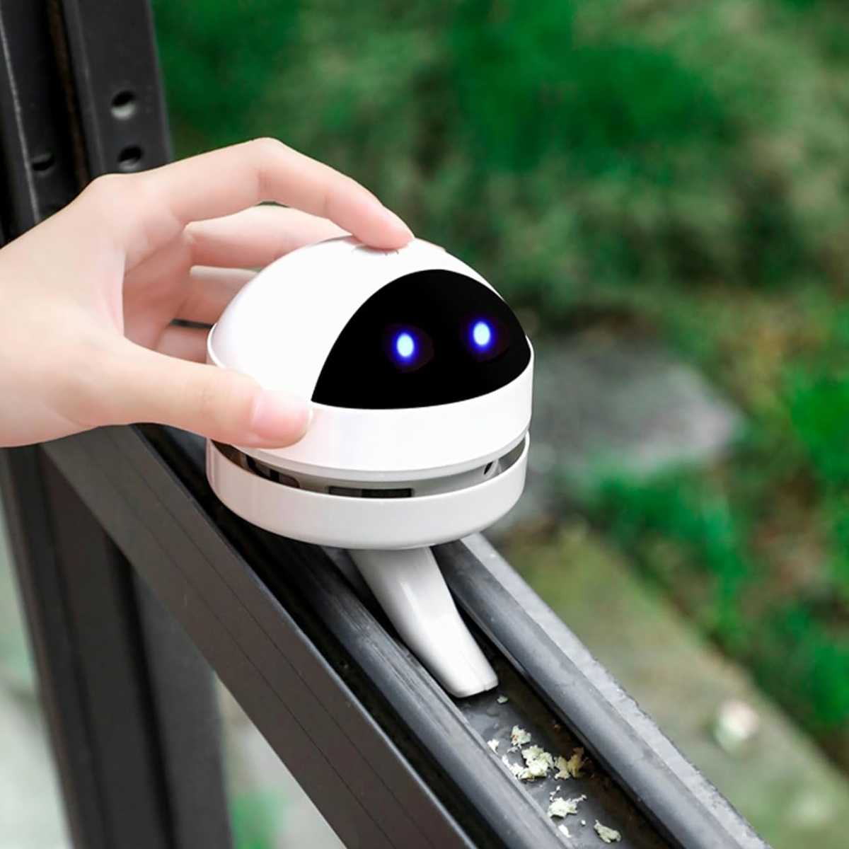 Eine Hand hält einen weißen Roboterreiniger auf einer Fensterschiene. Der Roboter hat blaue Augen. Im Hintergrund ist grünes Laub zu sehen.