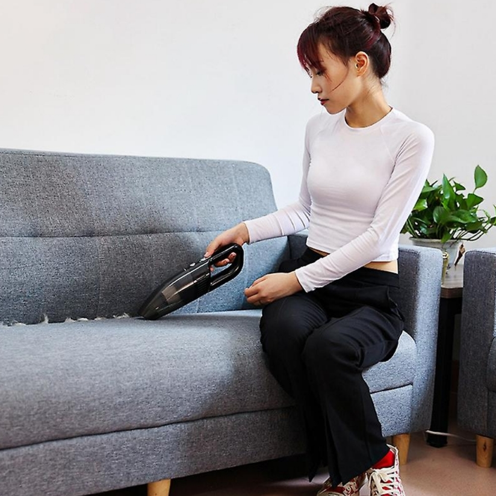 Eine Frau saugt mit einem schwarzen Handstaubsauger ein graues Sofa ab. Sie trägt ein weißes Oberteil, eine schwarze Hose. Pflanzen im Hintergrund.