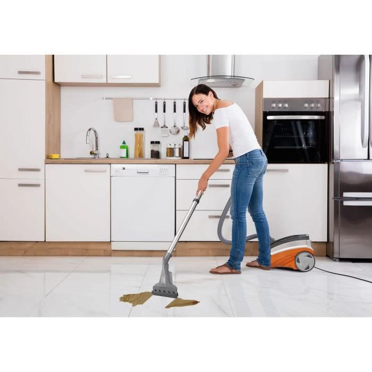 Eine Frau saugt einen Küchenboden. Sie trägt Jeans und ein weißes Hemd. Ein Staubsauger mit orange-grauem Gehäuse ist sichtbar.