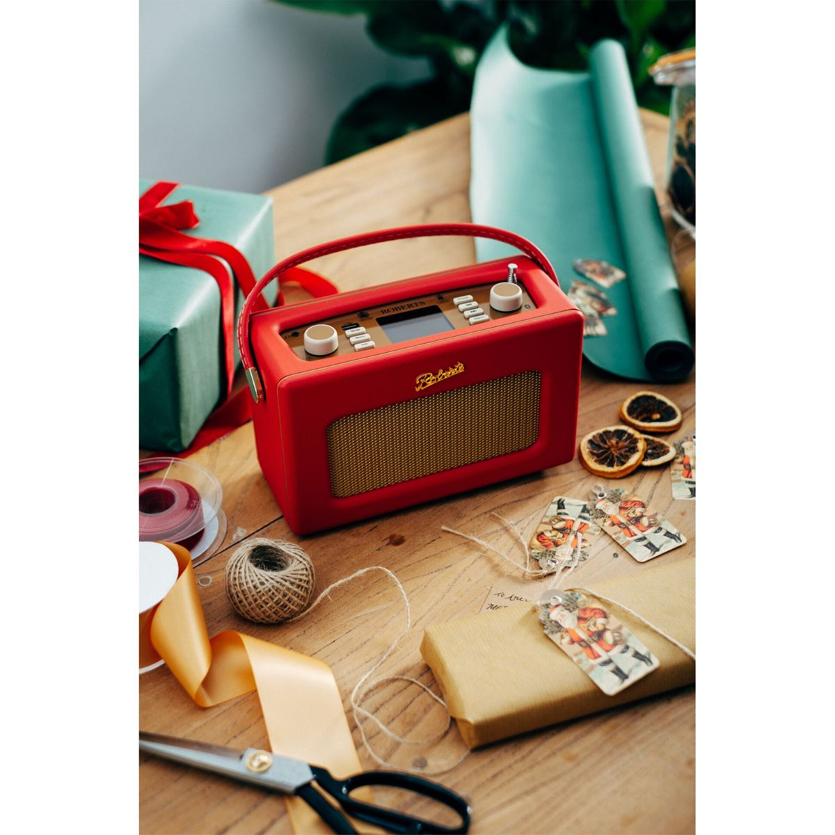 Una radio rossa con altoparlante dorato su un tavolo di legno, con regali, nastri, etichette e forbici. Tema natalizio.