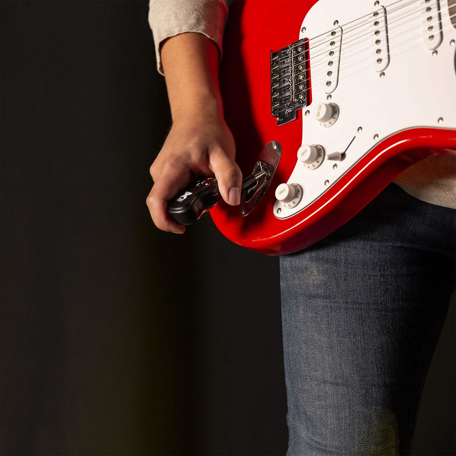 Una persona sostiene un dispositivo negro cerca de una guitarra eléctrica roja y blanca. La persona lleva camisa gris y vaqueros.
