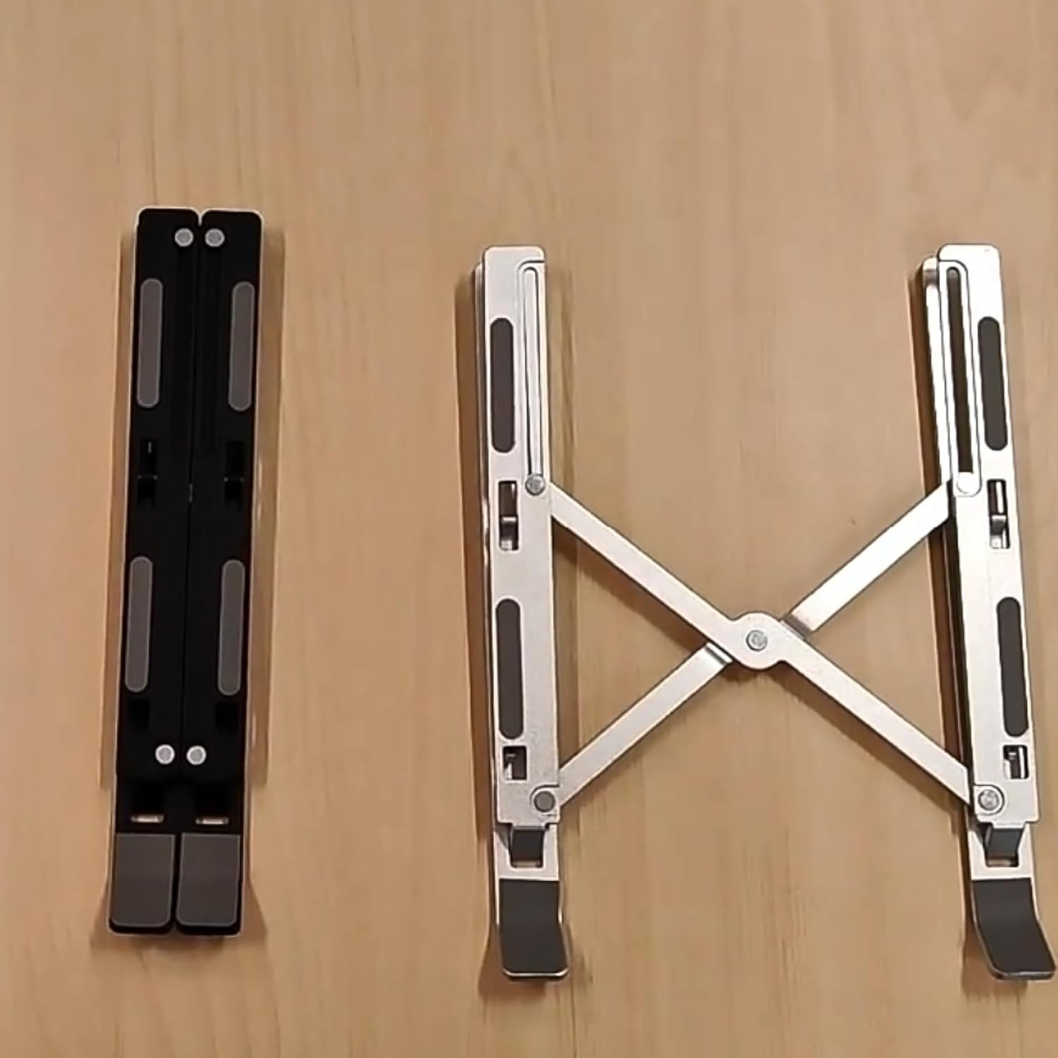 Zwei Laptop-Ständer. Einer schwarz, gefaltet. Der andere, silberfarben, in X-Form geöffnet. Beide auf einer Holzoberfläche.