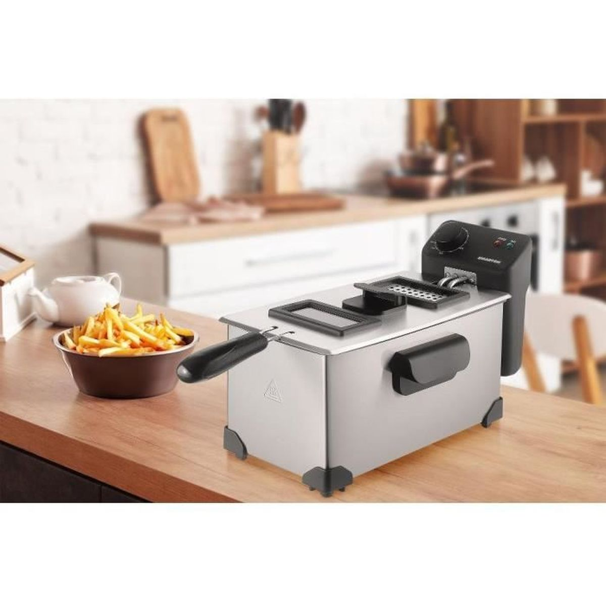 Una friggitrice in acciaio inossidabile su un tavolo di legno, con una ciotola di patatine fritte. Sfondo della cucina con utensili e decorazioni.