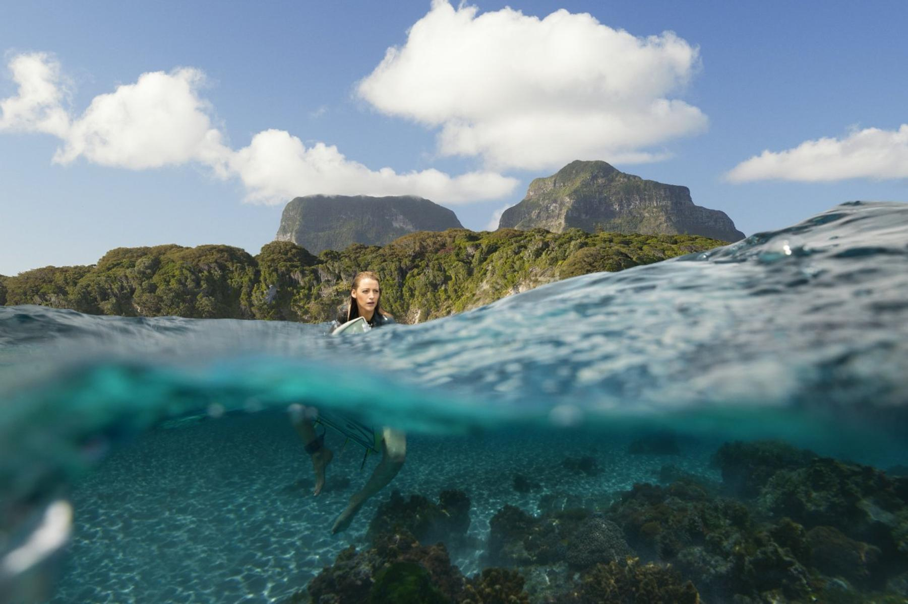 Une femme nage dans une eau claire, montagnes en arrière-plan. Au-dessus, ciel bleu avec des nuages. Fond marin rocheux visible.