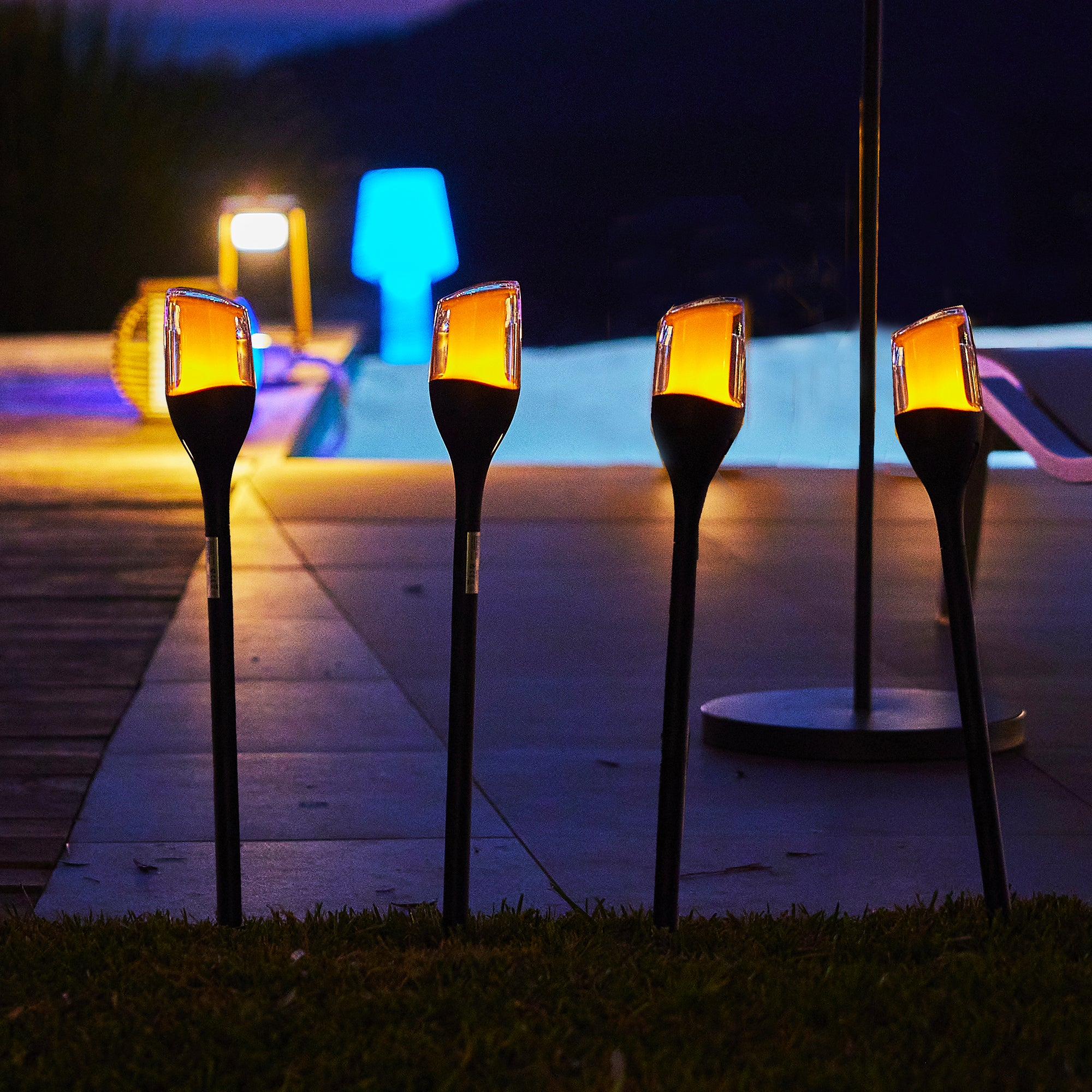 Vier beleuchtete Fackeln mit orangefarbenem Licht. Im Hintergrund eine blaue und eine gelbe Lampe.