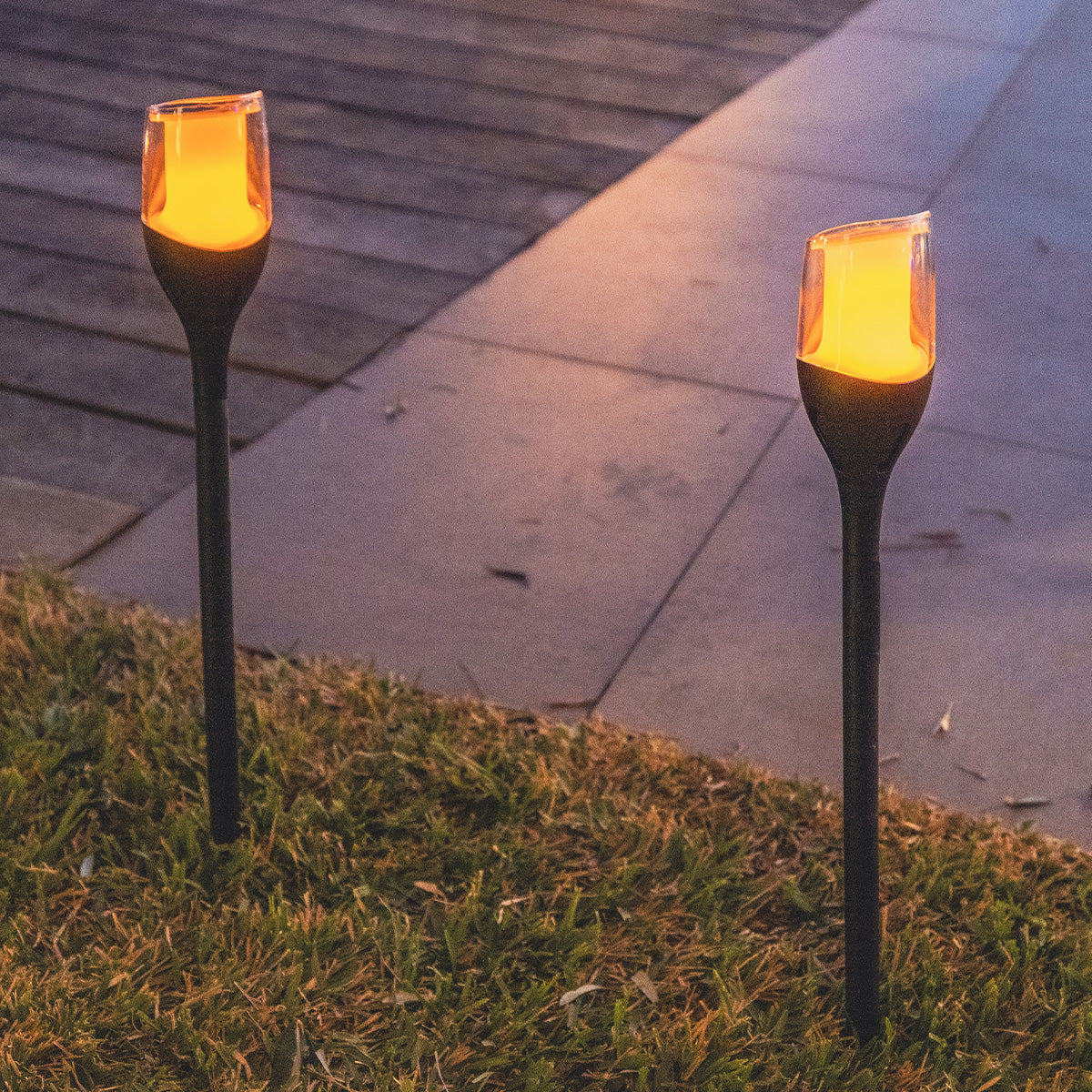 Zwei Außenlampen mit orangefarbenem Licht stehen im Gras. Im Hintergrund ist ein Weg.
