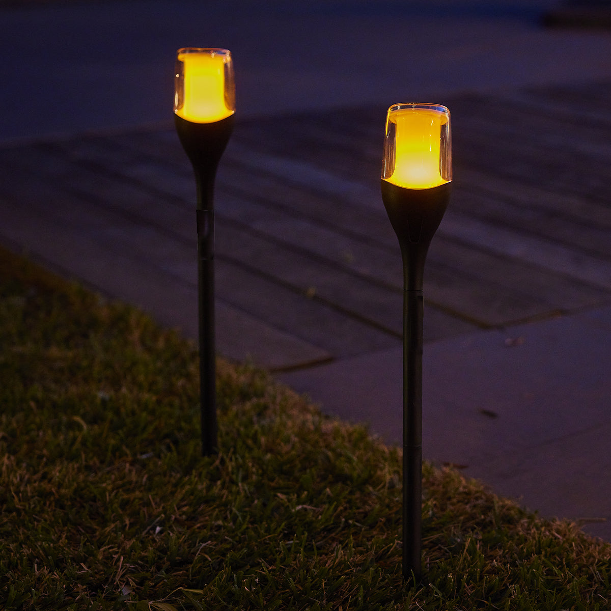 Zwei beleuchtete Gartenlichter mit gelbem Schein auf Gras und Holzweg bei Dämmerung.