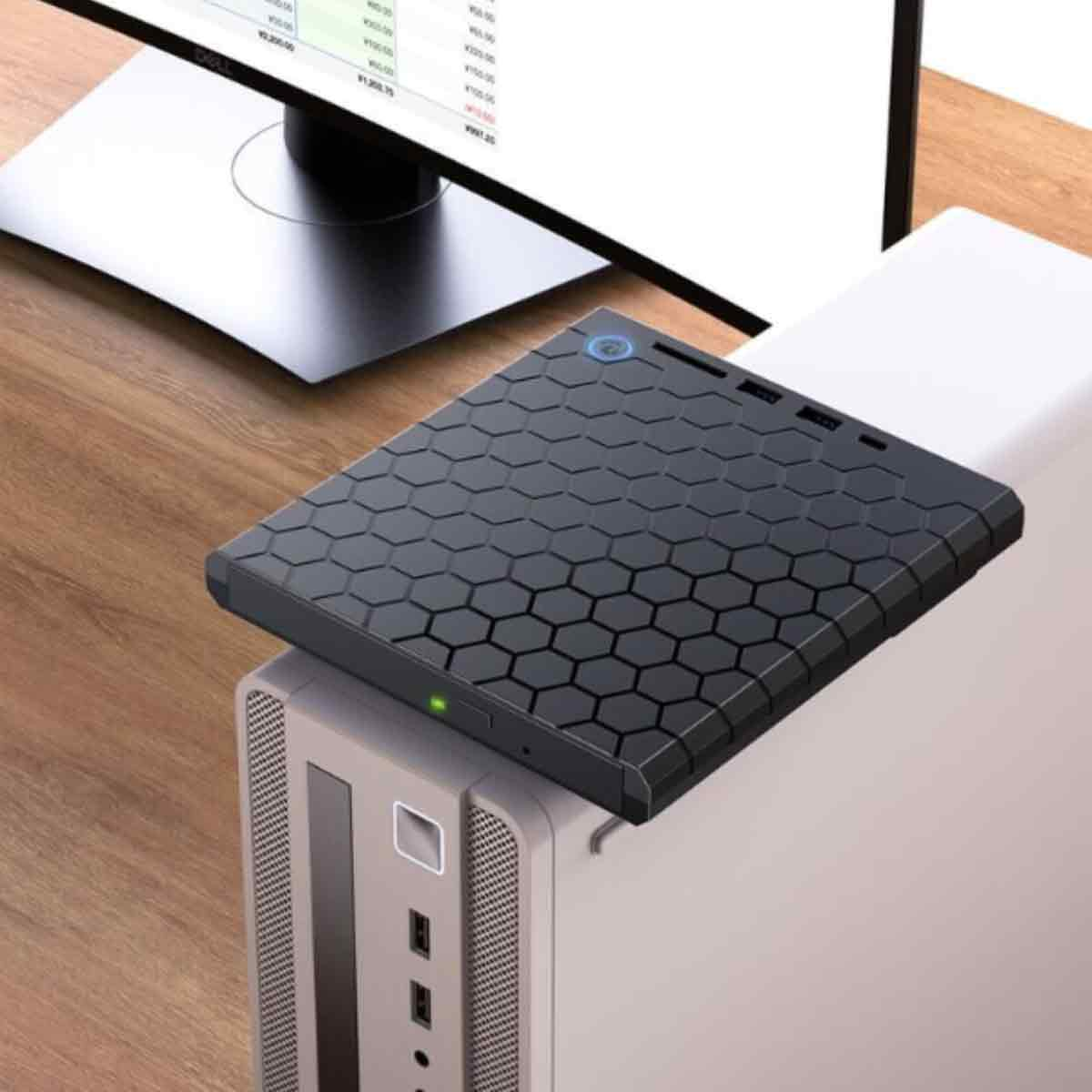 Ein schwarzes Gerät mit Wabenmuster liegt auf einem Computerturm. Ein Monitor zeigt Daten im Hintergrund auf einer Holzoberfläche.