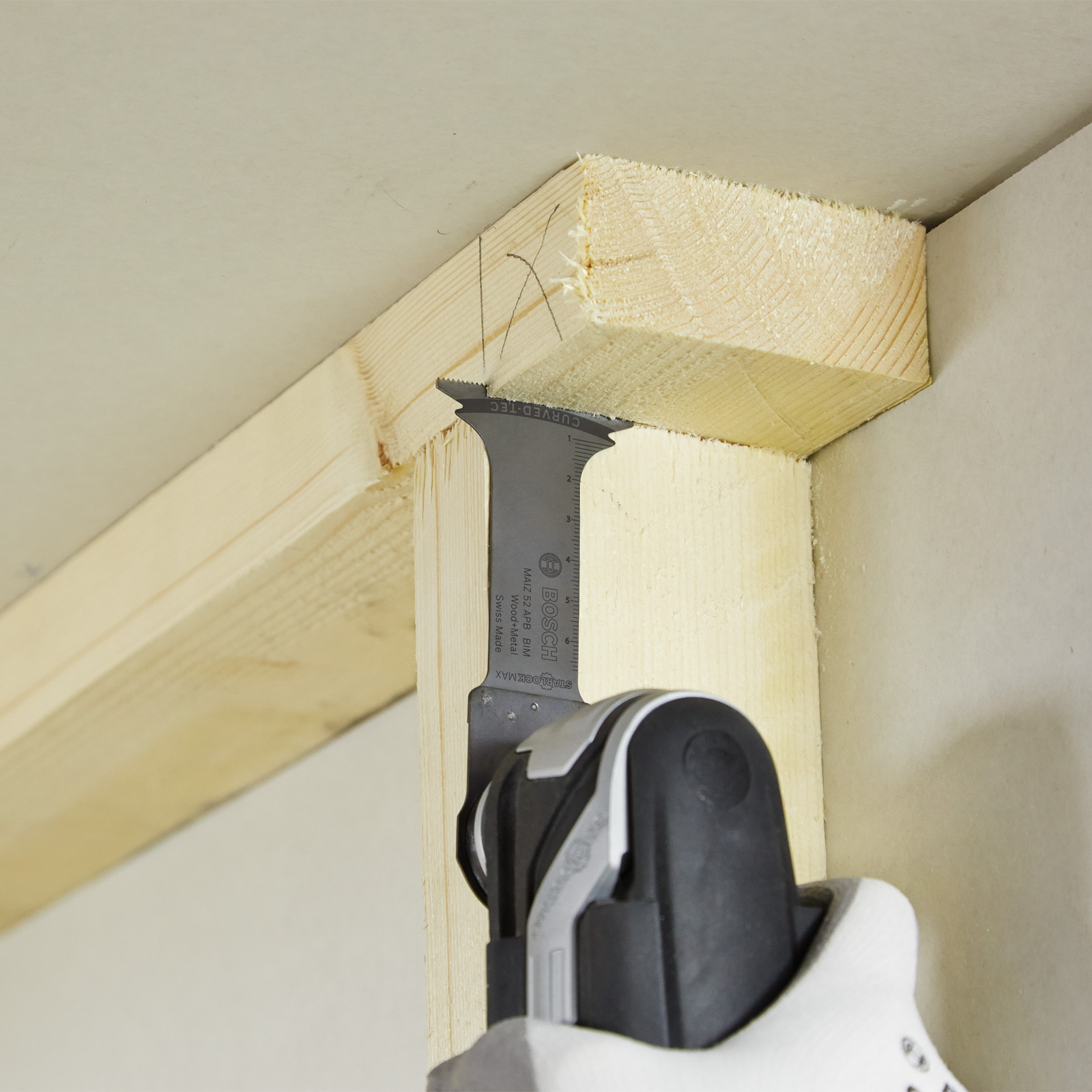 Una lama di sega taglia un telaio di legno. Una mano guantata di bianco tiene l'utensile. Lo sfondo è un muro bianco.