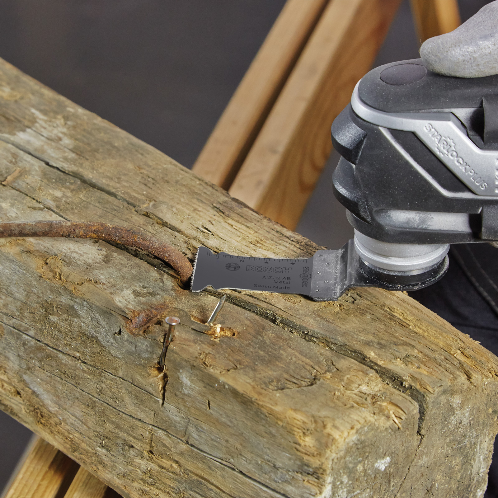 Una lama taglia una trave di legno. Un chiodo arrugginito e una sega metallica sono in primo piano. Lo sfondo mostra una struttura in legno.
