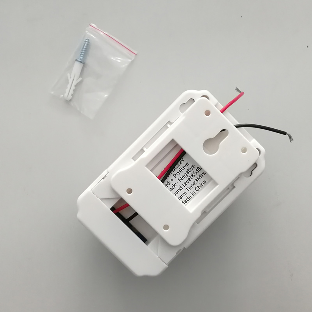 Ein weißes Sicherheitsgerät mit Kabeln und einer Plastiktüte mit einer Schraube und einem Wanddübel, auf einer hellgrauen Oberfläche.