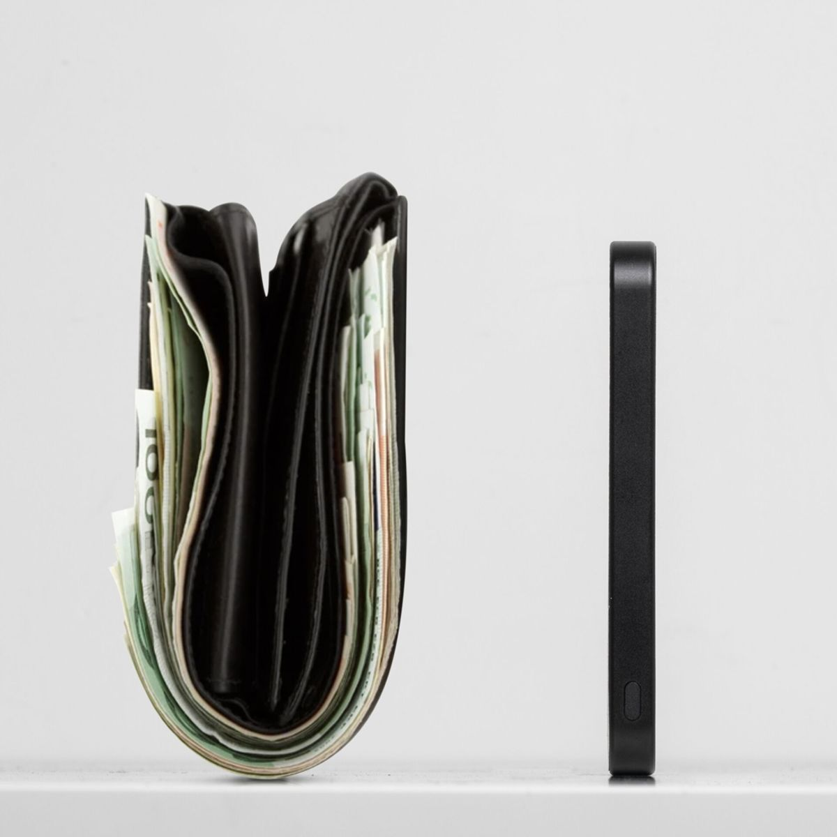 Eine schwarze Geldbörse voller Geld steht neben einem schwarzen Smartphone, beides auf einer weißen Oberfläche vor weißem Hintergrund.
