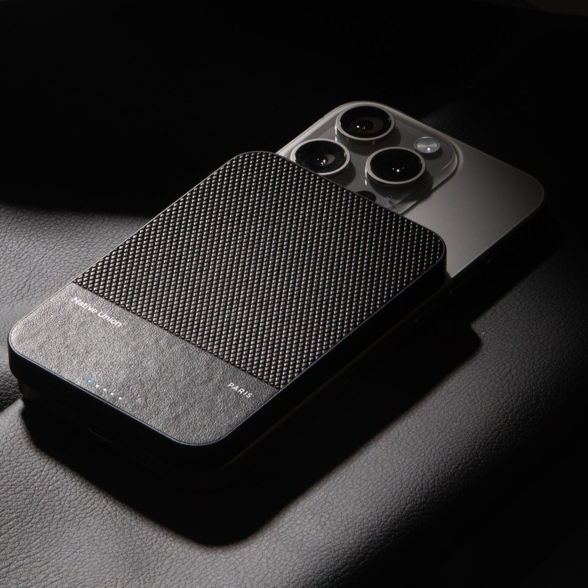 Ein Telefon mit einer schwarzen, strukturierten Powerbank, die auf schwarzem Leder liegt. Das Telefon hat drei Kameralinsen.