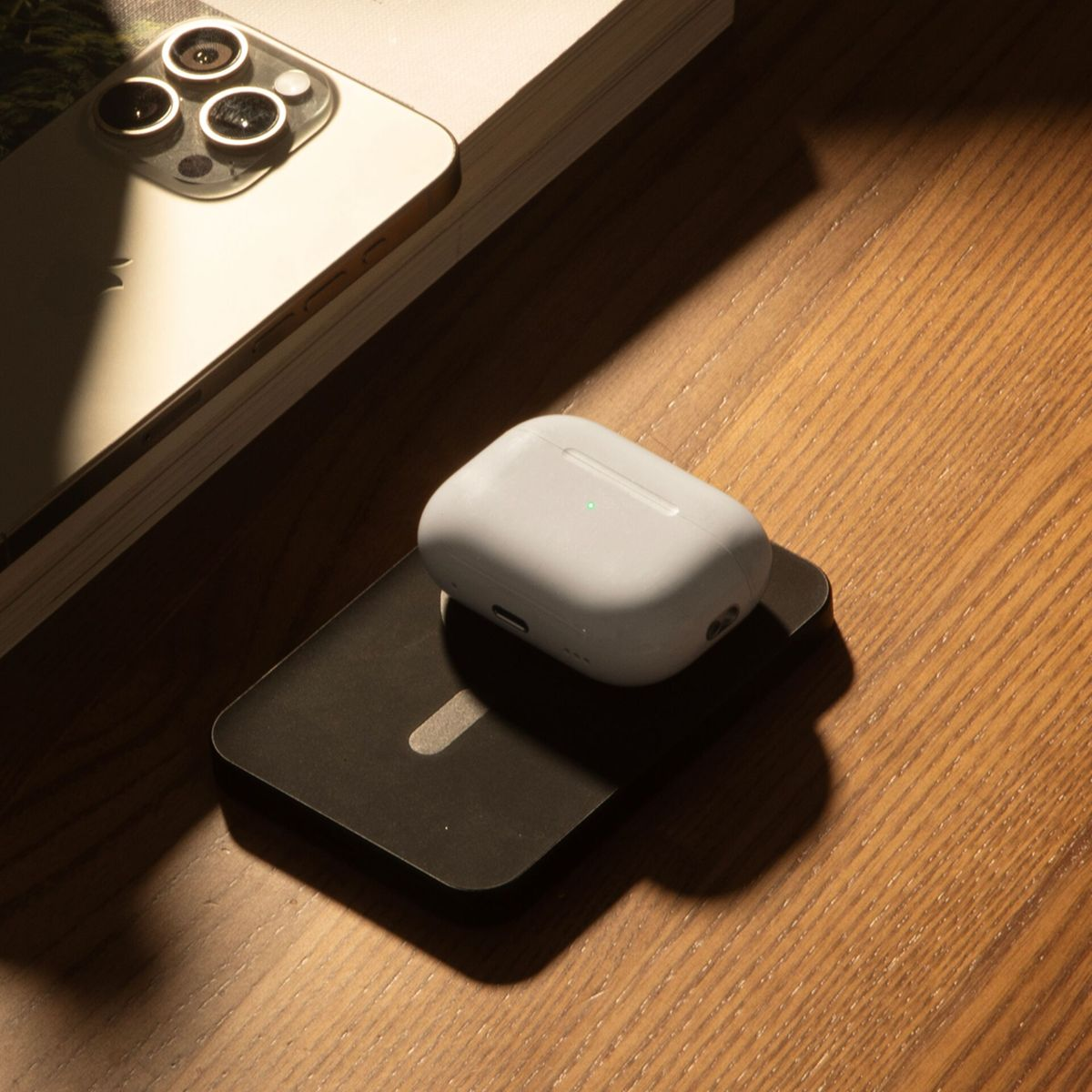 Ein weißes AirPods Pro-Etui liegt auf einem schwarzen Ladepad, neben einem Smartphone. Der Hintergrund ist eine Holzoberfläche.