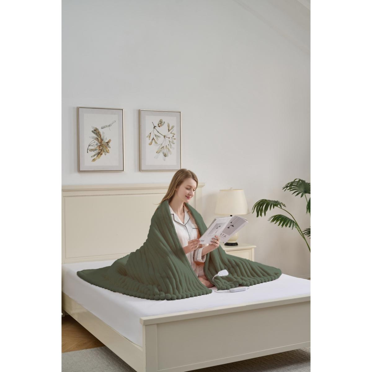 Eine Frau sitzt auf einem Bett, in eine grüne Decke gehüllt, und liest eine Zeitschrift. Eine Lampe, eine Pflanze und Bilder sind im Hintergrund.