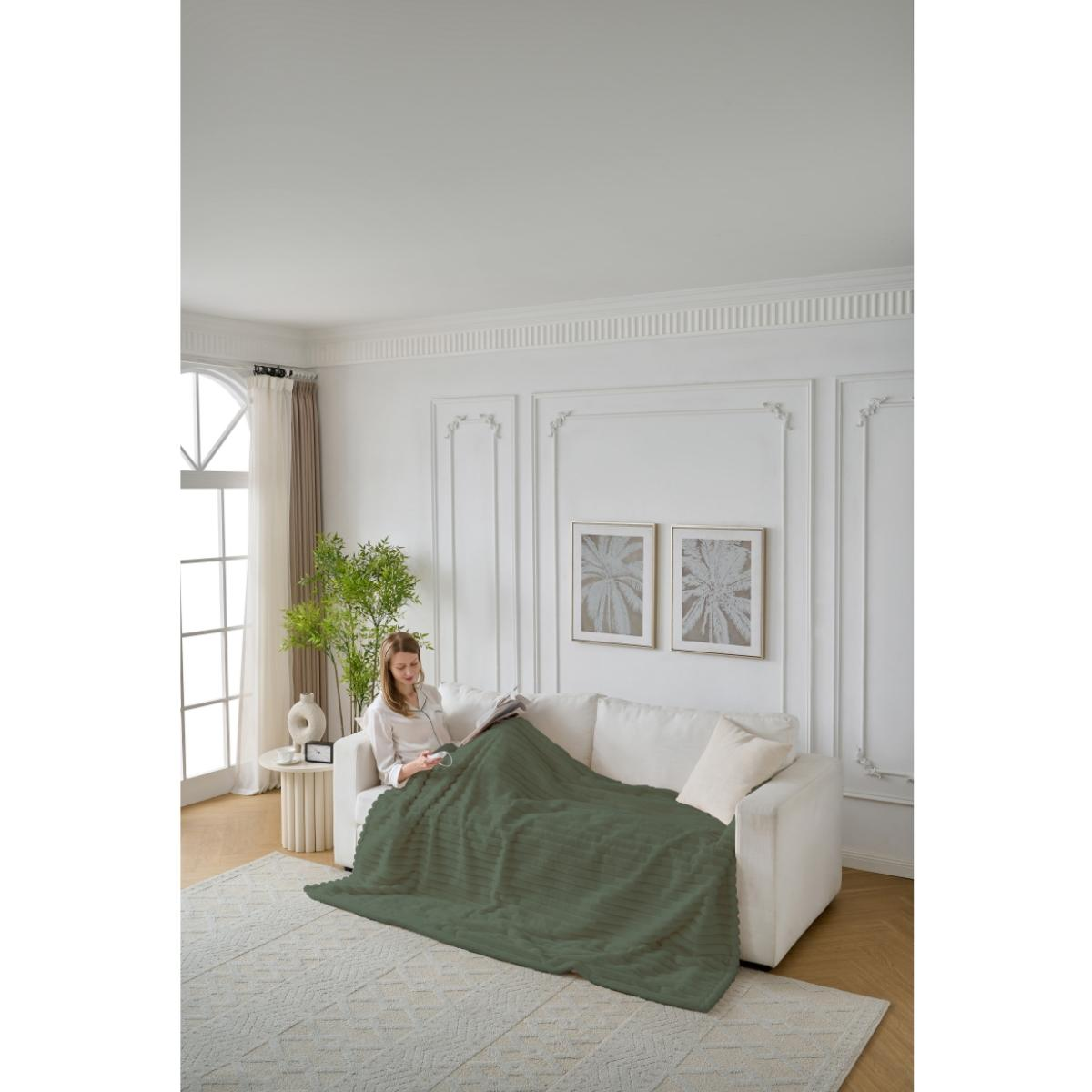 Frau auf weißem Sofa liest unter grüner Decke. Raum hat Teppich, Pflanze und Fenster.