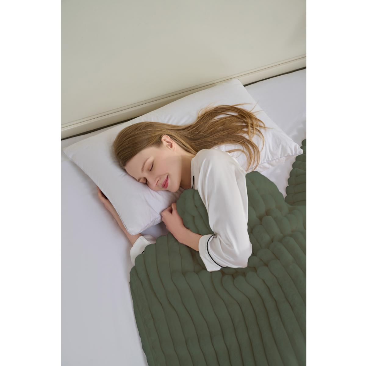 Eine Frau schläft im Bett, bedeckt von einer grünen Decke. Sie hat lange Haare und trägt ein weißes Hemd. Das Bett hat ein weißes Kissen.