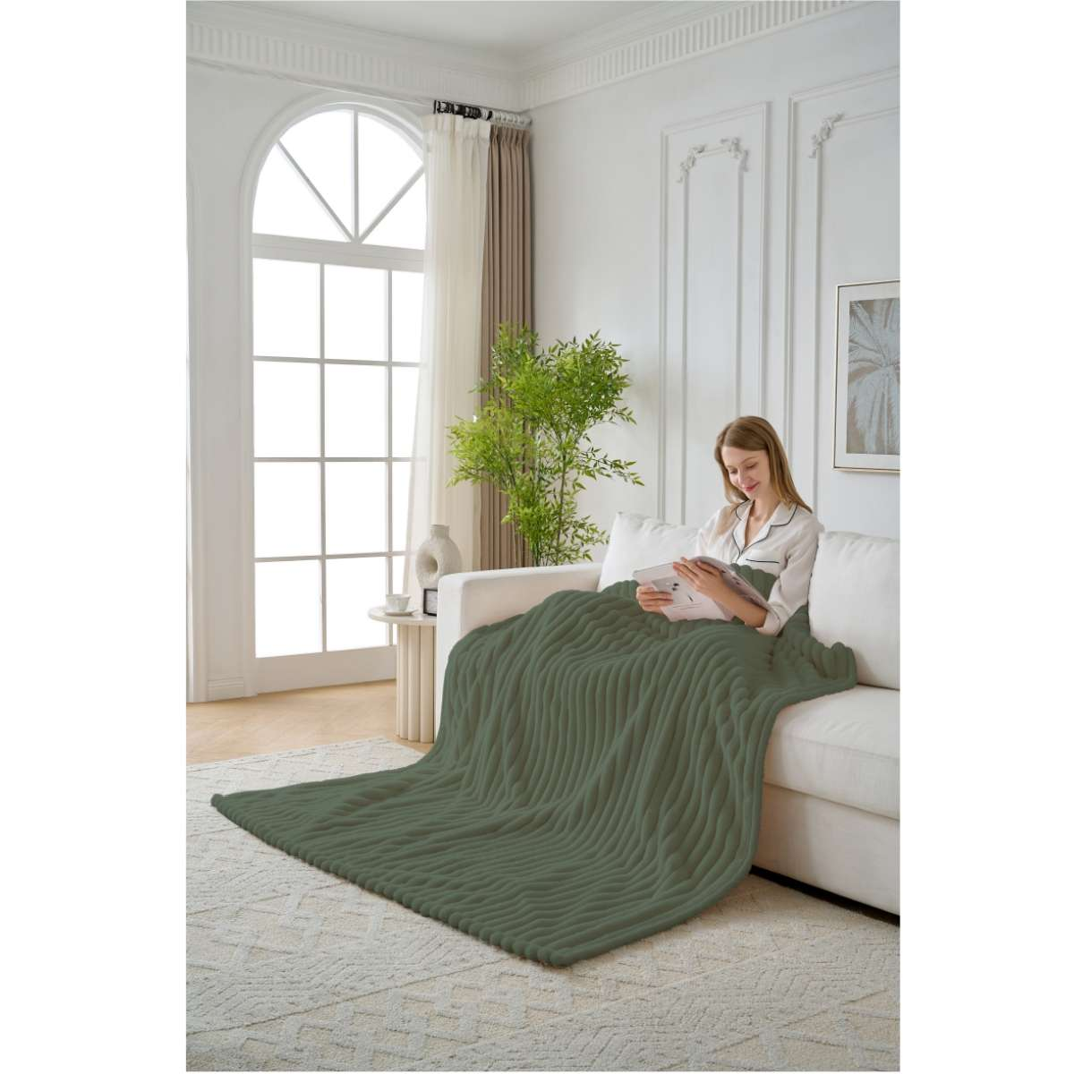Eine Frau sitzt auf einem weißen Sofa und liest unter einer olivgrünen Decke. Im Hintergrund ein großes Fenster und eine Pflanze.