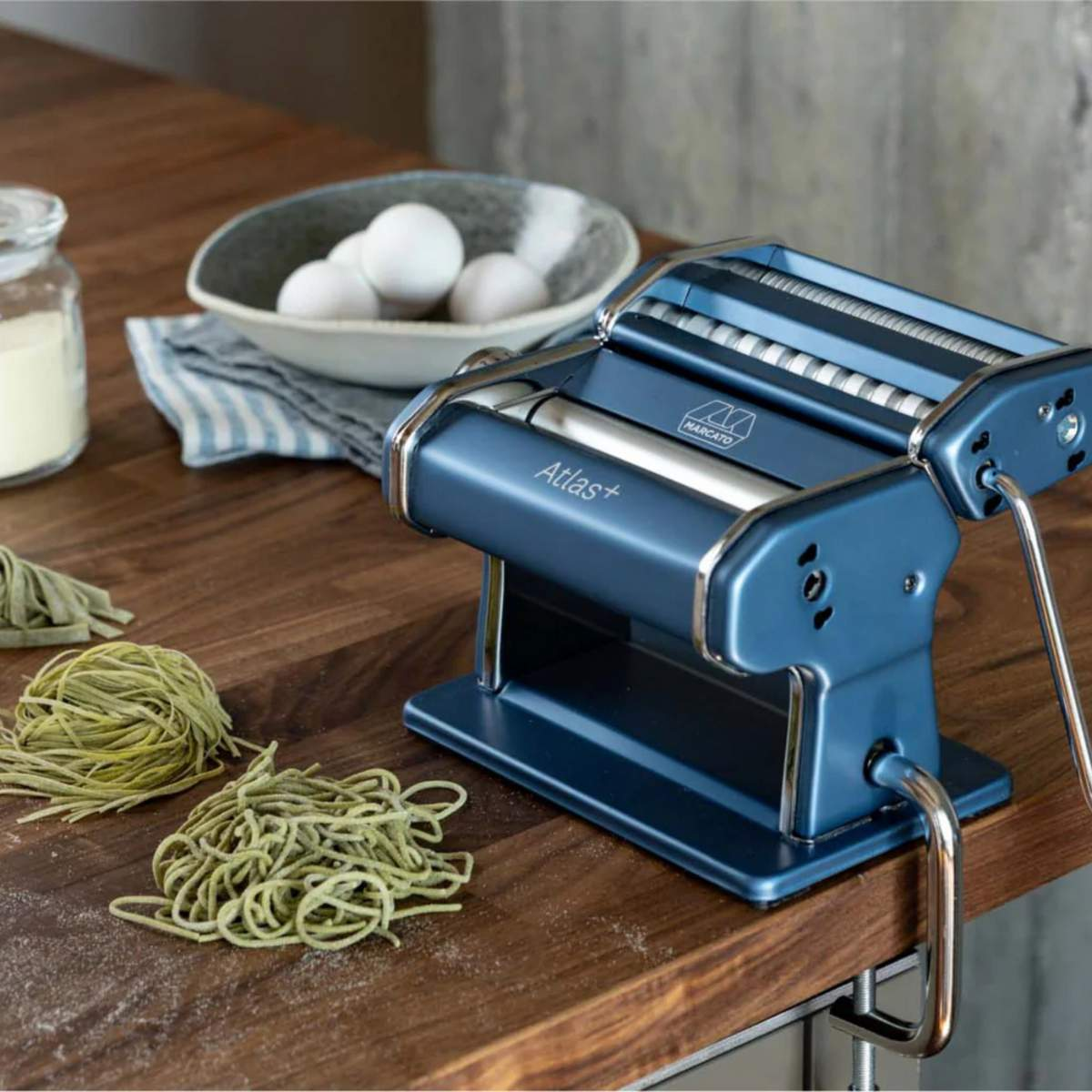 Blaue Pastamaschine auf Holzoberfläche mit Pasta, Eiern und Mehl. Die Maschine trägt die Aufschrift 'Atlas+'.
