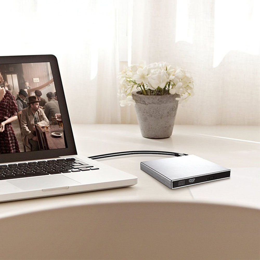 Laptop mit Filmszene, silbernes DVD-Laufwerk und Topf mit weißen Blumen auf weißem Tisch.