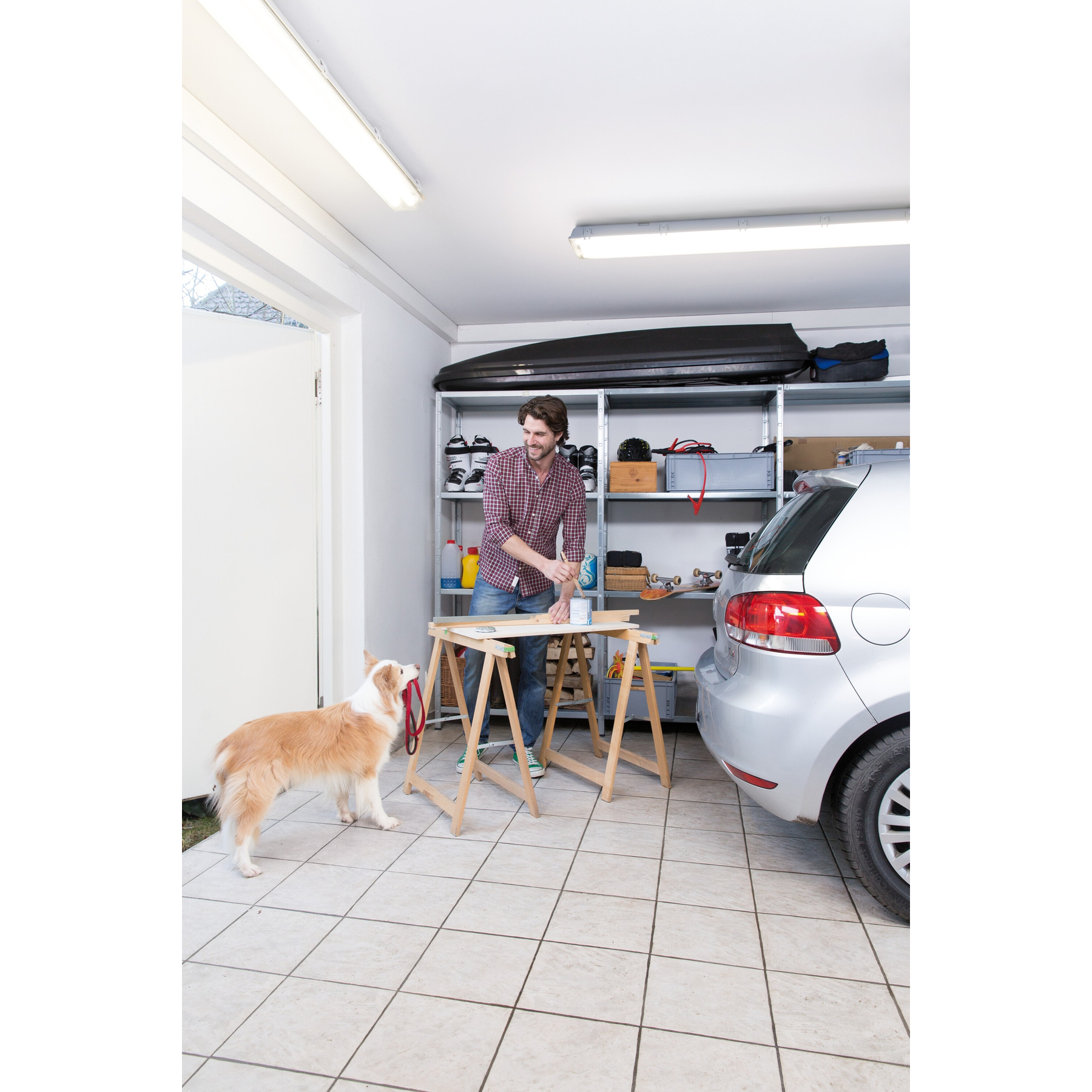 Mann malt Holz in einer Garage. Ein Hund mit Leine steht daneben. Im Hintergrund sind ein Auto und Regale zu sehen.