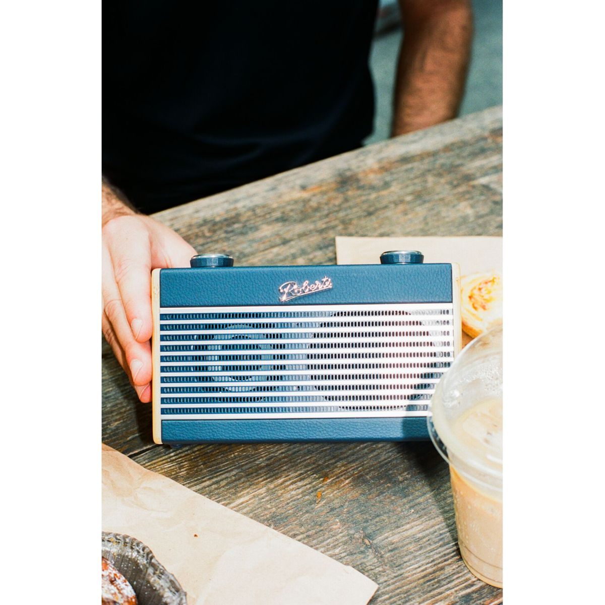 Eine Person hält ein blaues Roberts-Radio auf einem Holztisch. Kaffee und Gebäck sind in der Nähe.