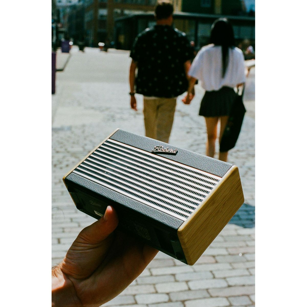 Eine Hand hält einen Vintage-Lautsprecher. Im Hintergrund gehen zwei Personen auf einer Kopfsteinpflasterstraße weg. Der Lautsprecher hat ein Holz- und Schwarzes Design.