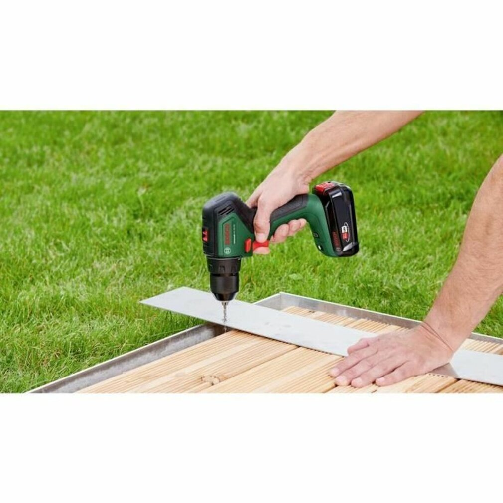 Eine Person benutzt einen grün-schwarzen Akkubohrer auf Holzplanken mit einer Metallführung. Grünes Gras im Hintergrund.