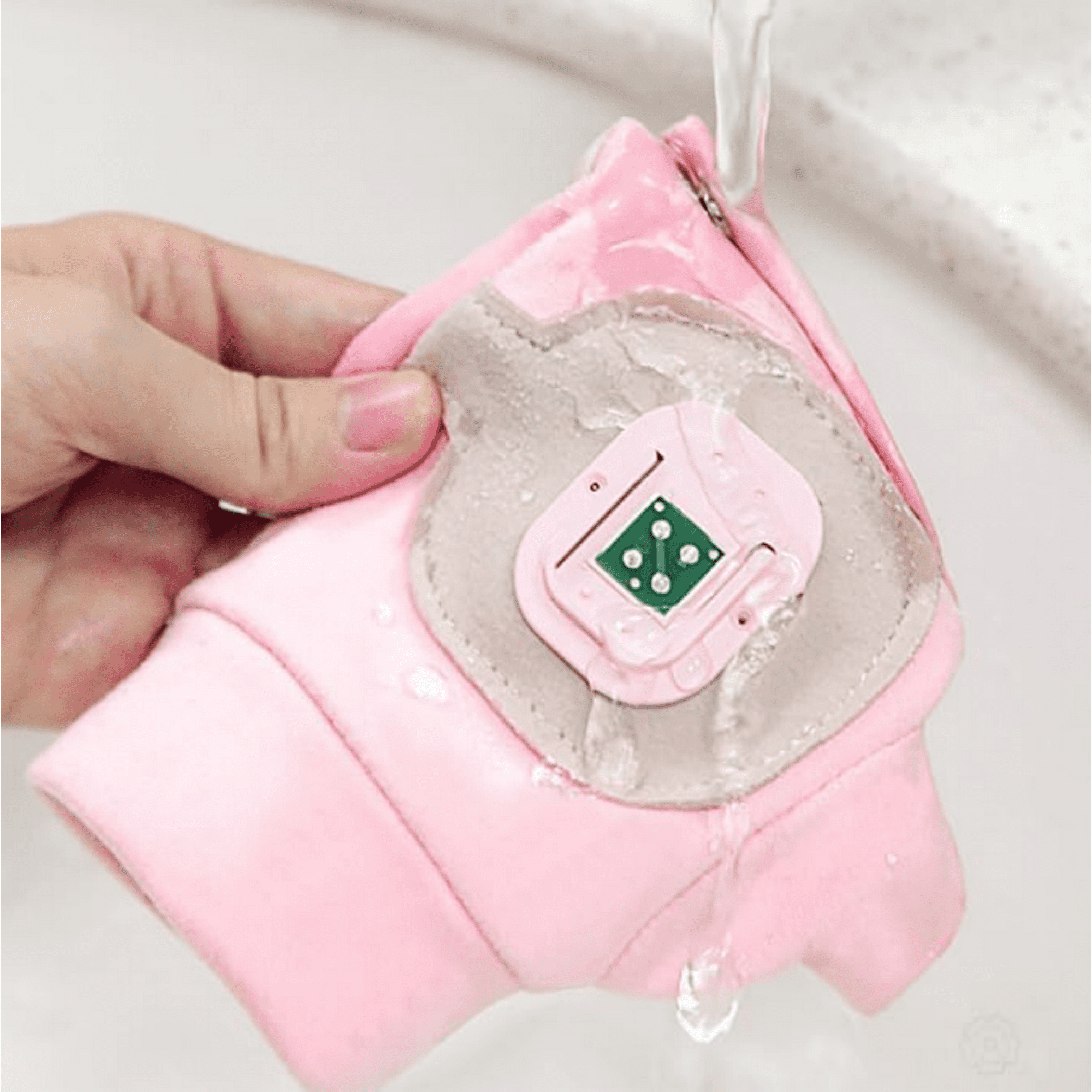 Eine Hand hält einen rosa Stoff mit einem grauen Aufnäher und einer grünen elektronischen Komponente. Wasser läuft darüber.