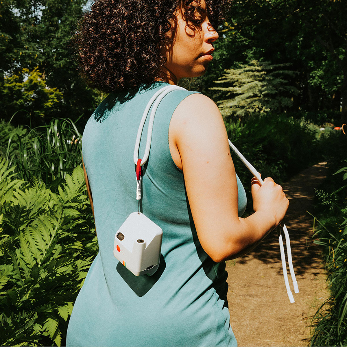 Una donna con i capelli ricci indossa un vestito verde acqua e porta una fotocamera bianca. Cammina su un sentiero in una foresta verde e frondosa.