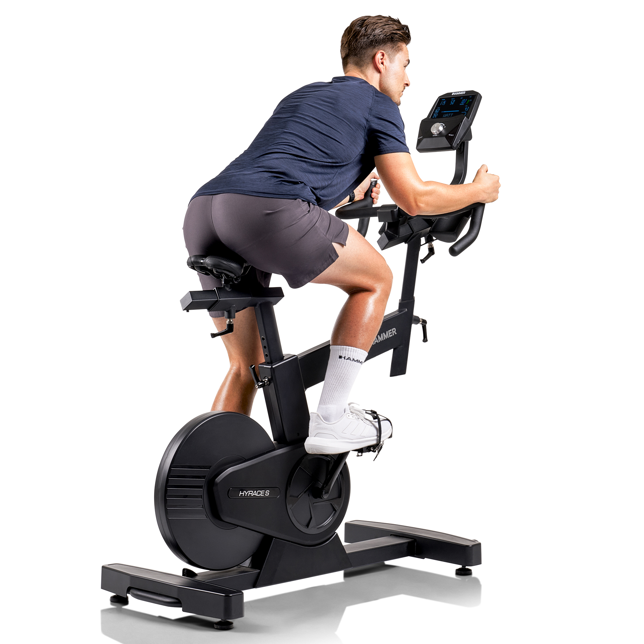 Ein Mann fährt auf einem schwarzen Heimtrainer. Er trägt ein blaues Hemd, graue Shorts und weiße Turnschuhe.