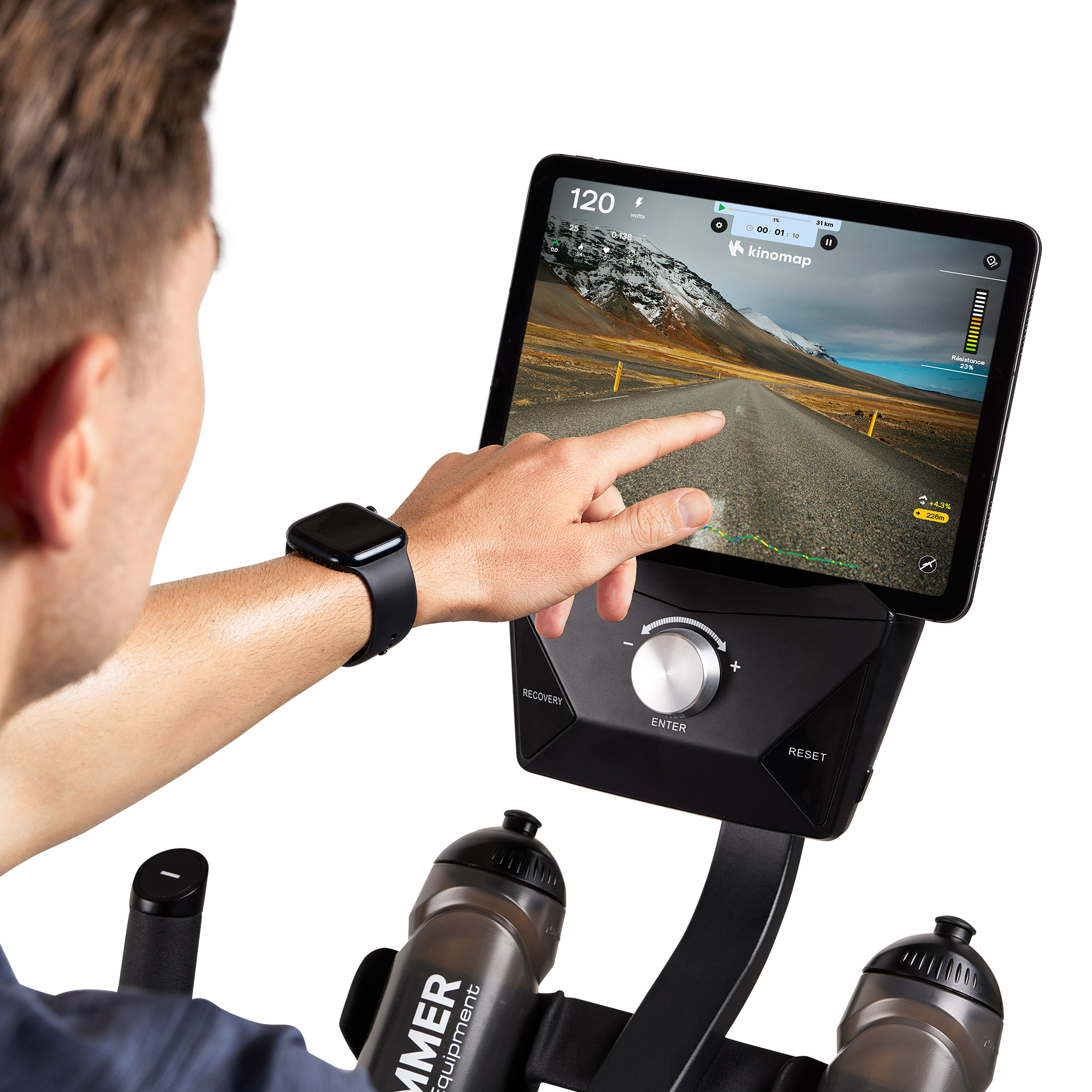 Eine Person benutzt ein Tablet auf einem Heimtrainer. Der Bildschirm zeigt eine Straße. Eine Hand berührt den Bildschirm.