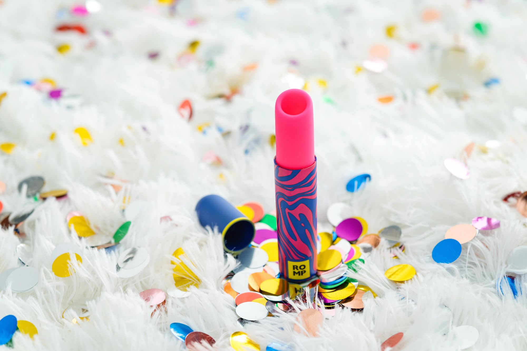 Un lápiz labial rosa está sobre una superficie blanca y esponjosa con confeti de colores. Un tubo azul yace al lado.