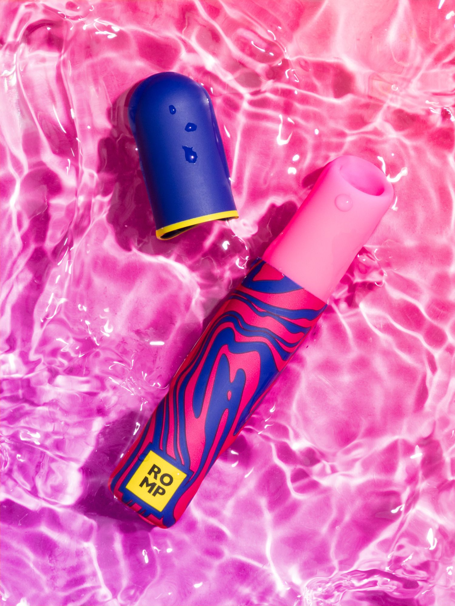 Un vibrador azul y rosa, con una tapa a juego, flota en agua rosa. El vibrador tiene un diseño estampado.