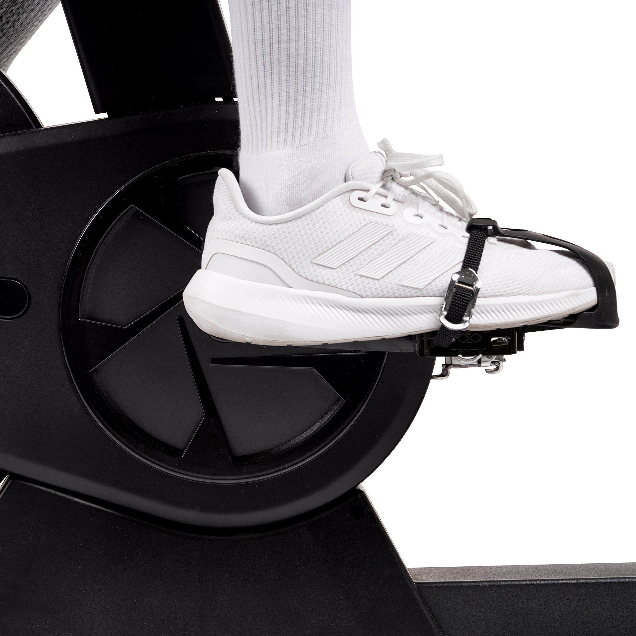 Fuß einer Person auf einem stationären Fahrradpedal. Weißer Schuh mit schwarzem Riemen. Schwarze Fahrradteile.
