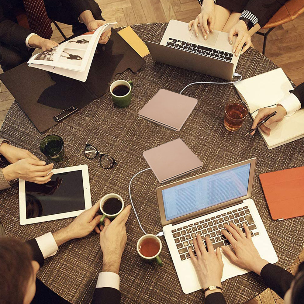 Menschen um einen runden Tisch mit Laptops, Kaffeetassen, Notizbüchern und einem Tablet. Hände tippen, lesen und halten Gegenstände.