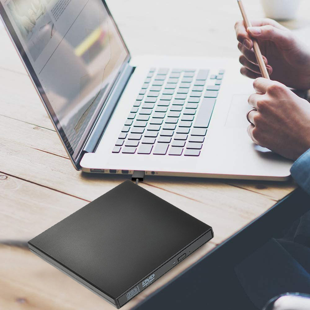 Auf einem Holztisch befindet sich ein Laptop mit einem schwarzen DVD-Laufwerk. Eine Person hält einen Bleistift über dem Laptop.