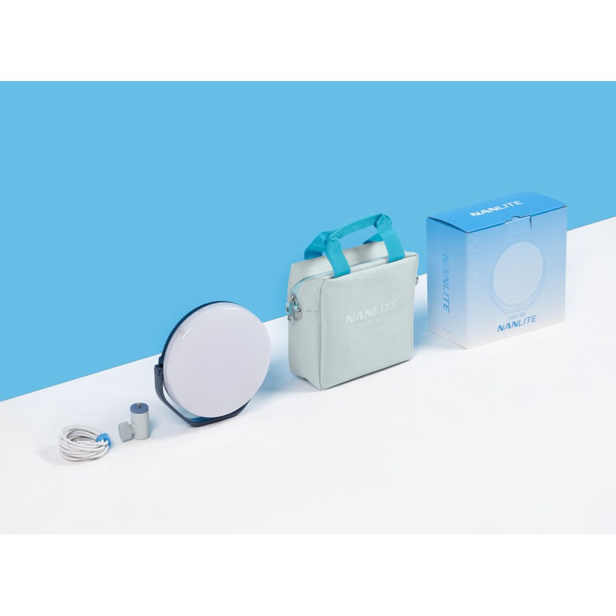 Una luce LED Nanlite bianca, una borsa e una scatola su una superficie bianca contro uno sfondo blu. Cavi e altri accessori sono presenti.