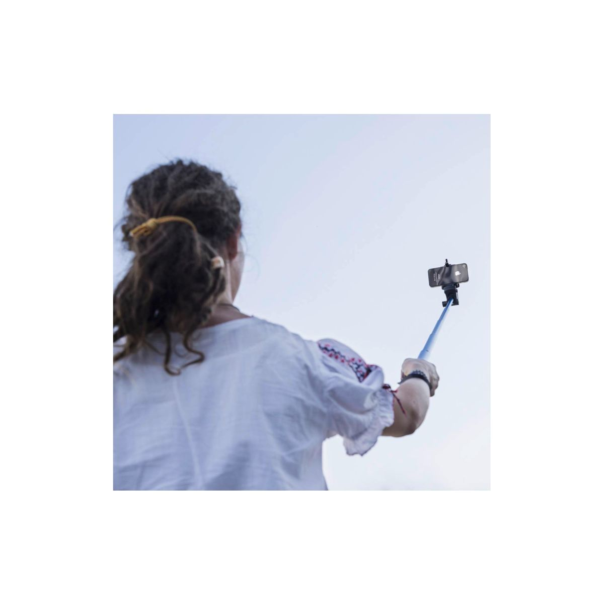 Eine Frau hält einen Selfie-Stick mit Telefon, blauer Himmel im Hintergrund. Sie trägt ein weißes Hemd mit roter Stickerei und hat dunkles Haar.