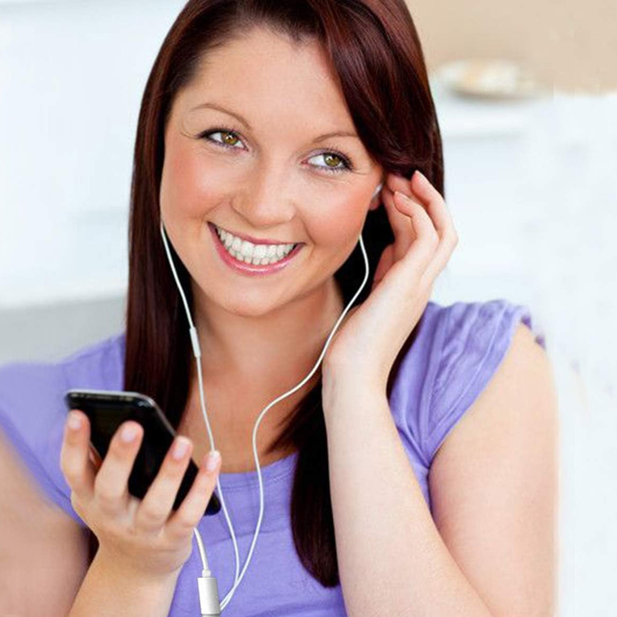 Eine lächelnde Frau mit roten Haaren hört Musik auf ihrem Handy mit weißen Kopfhörern. Sie trägt ein lila Hemd.