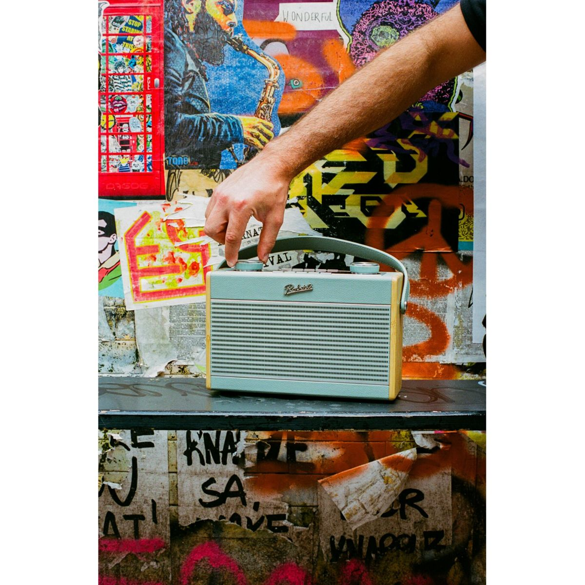 Eine Hand hält ein mintgrünes Radio mit Holzrahmen. Der Hintergrund ist eine mit Plakaten und Graffiti bedeckte Wand.