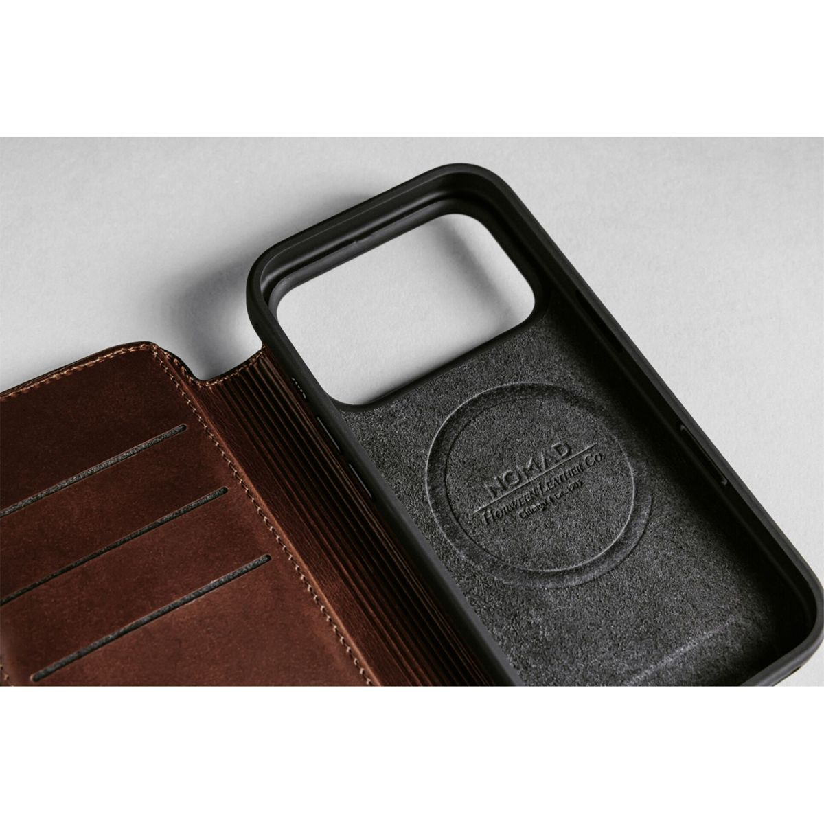 Nahaufnahme einer schwarzen Handyhülle und einer braunen Lederbrieftasche. Die Hülle hat ein NOMAD-Logo. Der Hintergrund ist grau.