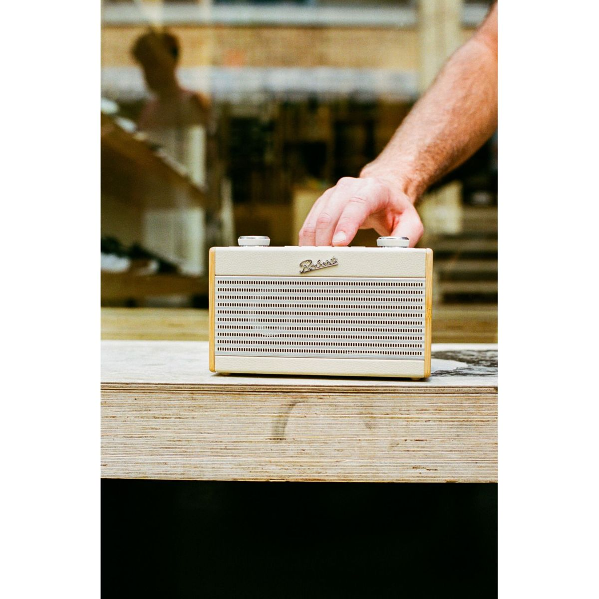 Ein cremefarbenes Roberts-Radio steht auf Holz. Eine Hand ruht darauf. Im Hintergrund sind eine verschwommene Person und ein Fenster zu sehen.